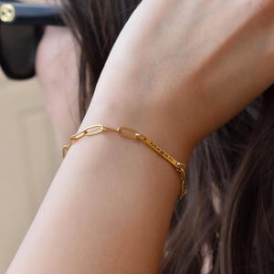 May include: A gold-colored chain bracelet with an elongated link design. The bracelet has a rectangular bar with the engraved name "Megan". The jewelry is worn on a person's wrist.