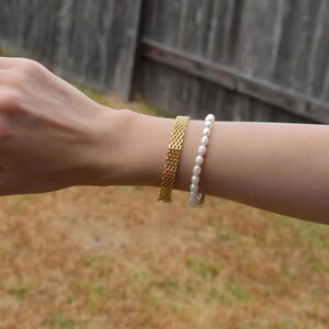 A person's hand wearing a gold bracelet and a white pearl bracelet is shown in the image.