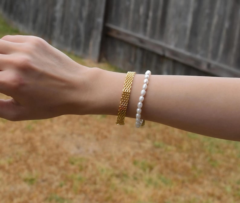 A person's hand wearing a gold bracelet and a white pearl bracelet is shown in the image.