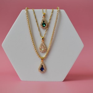 May include: Three gold-toned necklaces with teardrop pendants on a white display against a pink background. The pendants feature blue, clear, and green gemstones, each surrounded by smaller, clear stones. The necklaces vary in chain style.