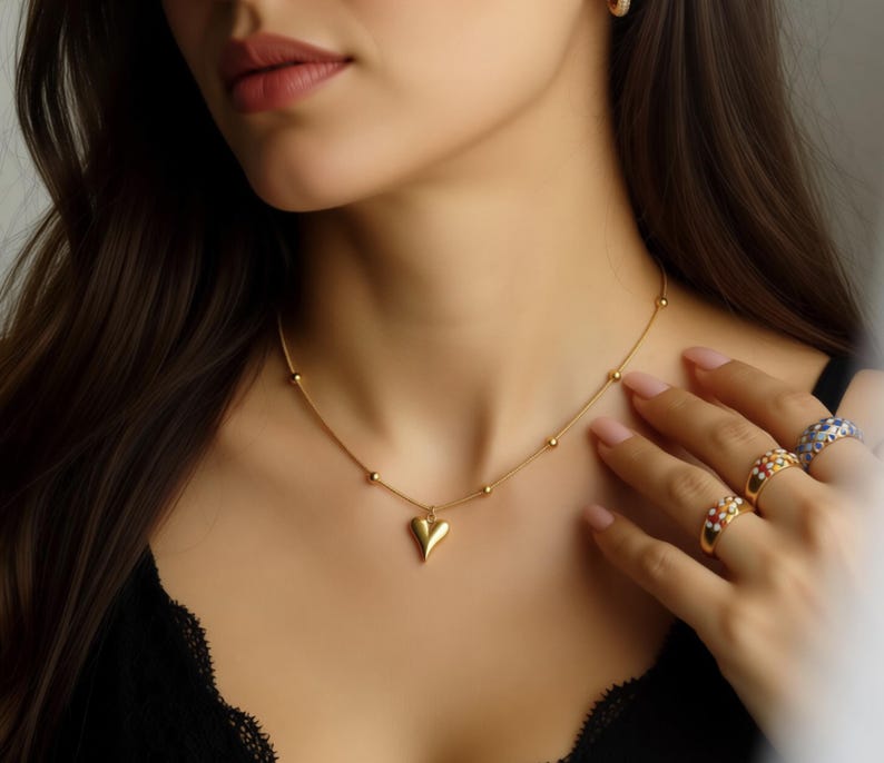 A close-up of a woman's face wearing a gold necklace with a heart-shaped pendant, and her hand resting on her shoulder.