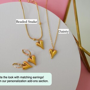 a collection of gold-colored heart-shaped pendants and earrings, including a necklace and a pair of earrings, arranged on a white surface against a pink background.