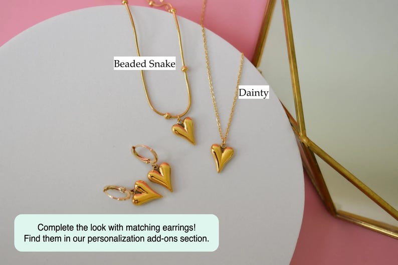 a collection of gold-colored heart-shaped pendants and earrings, including a necklace and a pair of earrings, arranged on a white surface against a pink background.