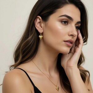 A woman with long brown hair wearing a black top and gold earrings with heart-shaped pendants, looking pensively to the side.