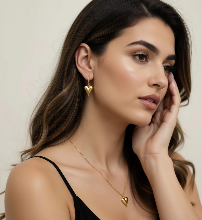 A woman with long brown hair wearing a black top and gold earrings with heart-shaped pendants, looking pensively to the side.