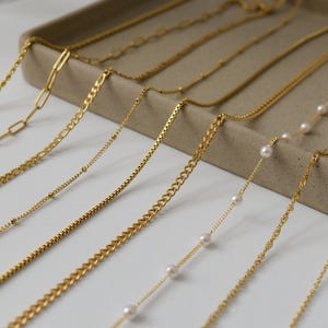 May include: A collection of gold necklaces with various chain styles, including link, curb, and beaded designs. Some necklaces feature small, white pearls. The jewelry is displayed on a neutral-toned tray.