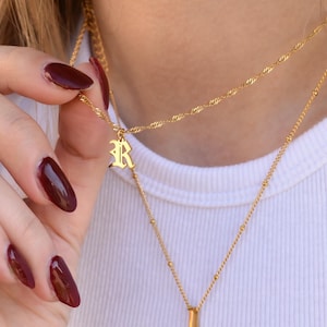 May include: Gold-colored layered necklaces. The top necklace has a twisted rope design, and the bottom necklace has small beads. The top necklace has a pendant with the letter "R". The person is wearing a white shirt and has burgundy nail polish.