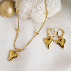 A gold necklace with a heart-shaped pendant and matching earrings are displayed on a white surface.