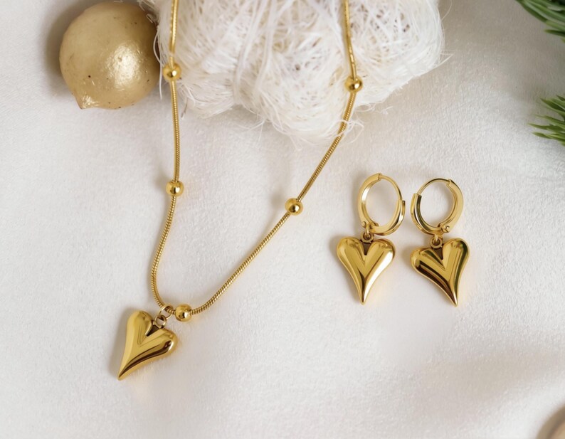 A gold necklace with a heart-shaped pendant and matching earrings are displayed on a white surface.