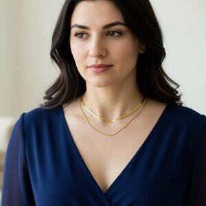 A woman with long dark hair wearing a blue dress and a gold necklace, looking off to the side with a thoughtful expression.