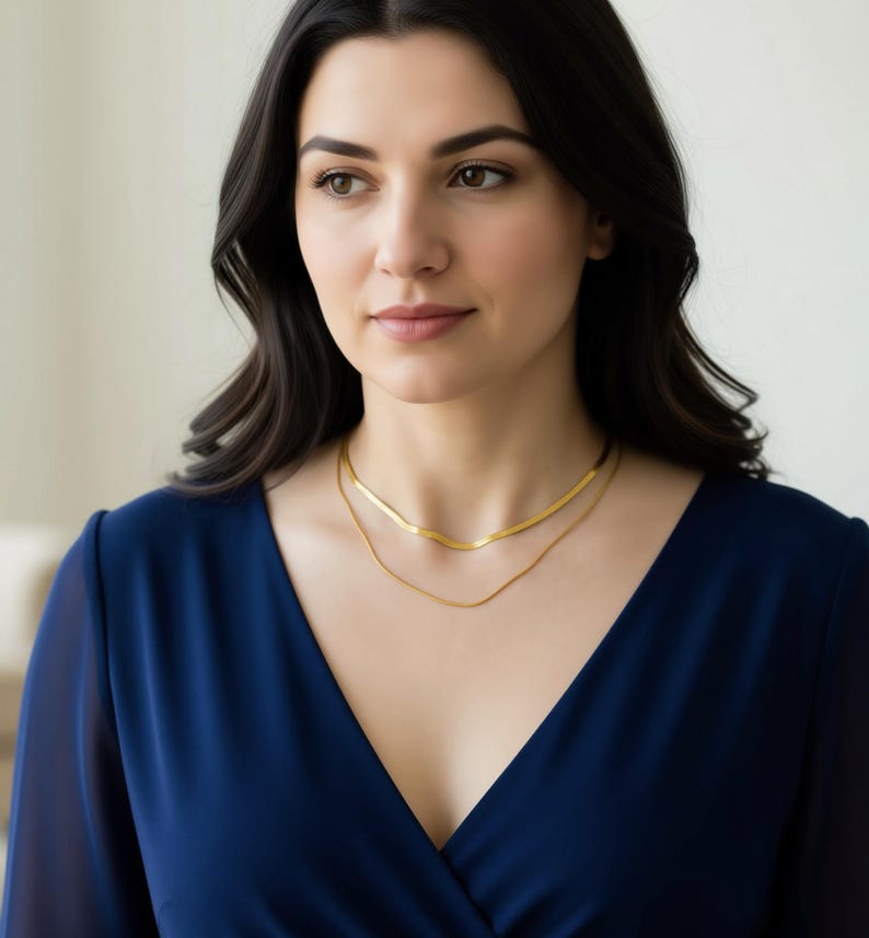 A woman with long dark hair wearing a blue dress and a gold necklace, looking off to the side with a thoughtful expression.