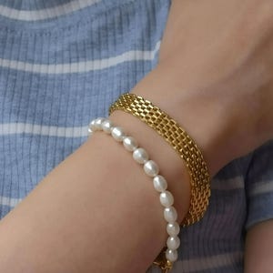 A close-up of a person's arm wearing a gold bracelet and a pearl bracelet, with their hair visible.