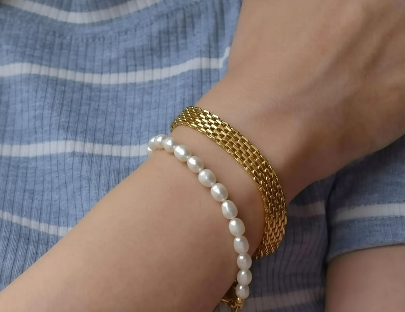 A close-up of a person's arm wearing a gold bracelet and a pearl bracelet, with their hair visible.