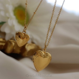 May include: Two gold-colored heart-shaped locket necklaces. The lockets hang from delicate chains. The larger locket has an ornate design on the front. The background includes a white flower and a gold-colored surface.