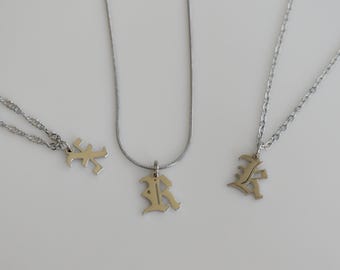 Op de afbeelding: Drie zilverkleurige kettingen met hangers in de vorm van de letter K in een gotisch lettertype. Elke hanger hangt aan een ander type zilverkleurige ketting, waaronder een gladde slangenketting en twee kettingen met gedraaide schakels.
