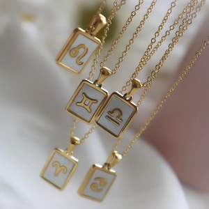 May include: Gold-toned necklaces with rectangular pendants featuring zodiac symbols. Each pendant has a white background and a gold border. The chains are delicate and the pendants are arranged at different lengths, creating a layered effect.