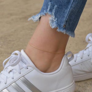 A close-up view of a person's ankle wearing white sneakers and a gold ankle bracelet.