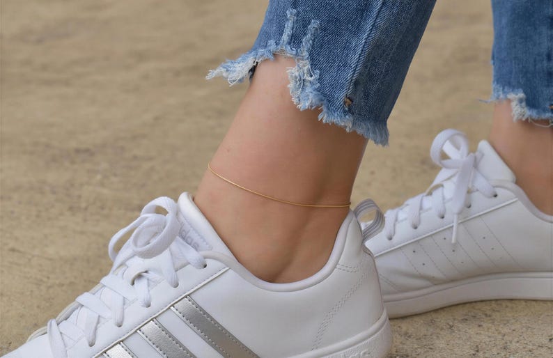 A close-up view of a person's ankle wearing white sneakers and a gold ankle bracelet.