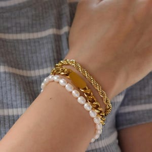 A close-up of a person's wrist wearing three bracelets: a gold chain bracelet, a pearl bracelet, and a gold tag bracelet.