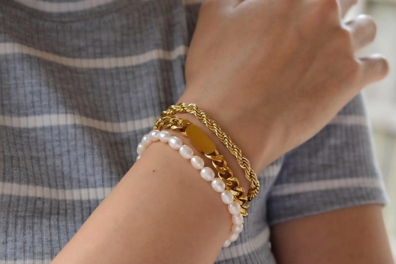 A close-up of a person's wrist wearing three bracelets: a gold chain bracelet, a pearl bracelet, and a gold tag bracelet.