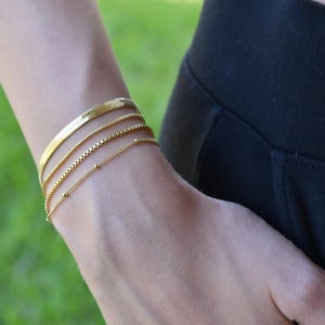 May include: A set of four gold-toned bracelets worn on the wrist. The collection includes a wide flat herringbone chain, a thin smooth snake chain, a square box chain, and a delicate cable chain featuring small evenly spaced round metal beads.