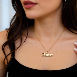 May include: A gold-colored necklace with the name "Palmer" in a serif font. The pendant hangs from a delicate chain. The person in the image is wearing a black top.