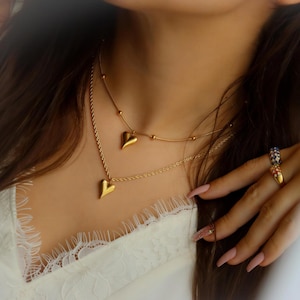 A close-up of a woman's neck and chest area, featuring a necklace with two heart-shaped pendants and a ring on her finger.