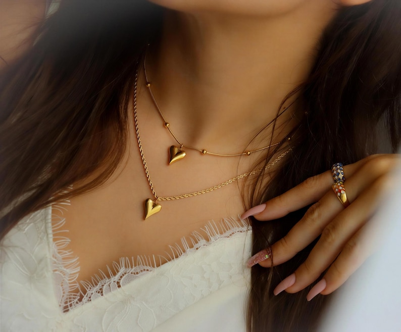 A close-up of a woman's neck and chest area, featuring a necklace with two heart-shaped pendants and a ring on her finger.