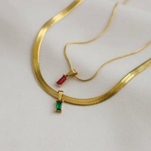 May include: Two gold-toned necklaces. One is a flat herringbone chain with a rectangular green pendant. The other is a thin snake chain with a smaller rectangular red pendant. Both pendants feature a gold-toned metal setting and a polished finish.