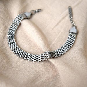 A silver metal bracelet with a twisted, braided design is lying on a beige fabric surface.