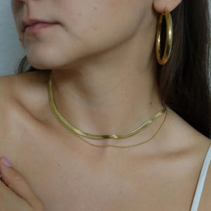 A close-up of a woman's neck and chest area, featuring a gold necklace and hoop earrings.