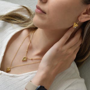 A close-up of a woman's face wearing a necklace with heart-shaped pendants and a pair of gold earrings.