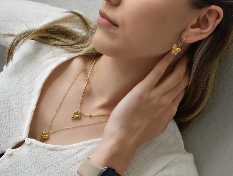 A close-up of a woman's face wearing a necklace with heart-shaped pendants and a pair of gold earrings.