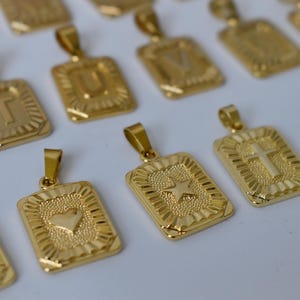 May include: A collection of rectangular gold-toned metal pendants arranged in rows. Each pendant features a textured center with a raised symbol, including letters of the alphabet, a heart, a star, and a cross, all with a polished border.