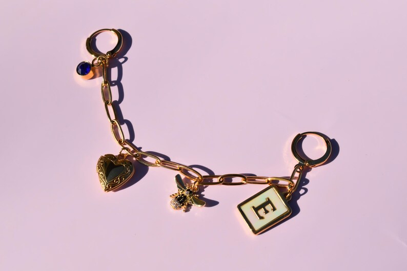 A gold-colored charm bracelet with various charms attached, including a heart-shaped charm, a bee charm, and a letter-shaped charm, all set against a pink background.