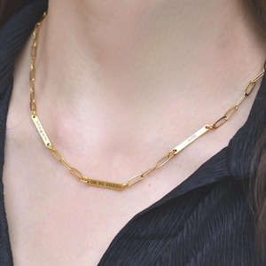 May include: A gold-colored chain necklace with three rectangular bars. The bars are engraved with the names "Megan" and "Aot", and the date "06.30.2022". The necklace is worn against a black top.