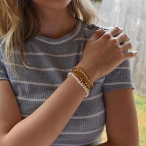 A close-up of a person's arm wearing a gray and white striped shirt, with a bracelet and multiple rings on their fingers.