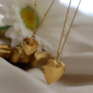 May include: Two gold-toned heart-shaped lockets suspended on delicate matching chains. Each locket features an intricate engraved scrollwork pattern along the edges, with a smooth, polished center surface. The text GoldBlonde is visible.