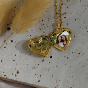 May include: A gold-toned heart-shaped locket necklace on a delicate chain. The locket is open, revealing the text I Love You engraved on the left side and a photograph of two smiling people on the right side, set against a textured stone surface.