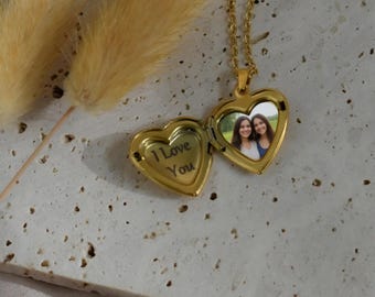 Puede incluir: Un collar con dije en forma de corazón dorado y cadena fina. El dije está abierto, mostrando el texto I Love You grabado en el lado izquierdo y una fotografía de dos personas sonrientes en el lado derecho, sobre una superficie de piedra.