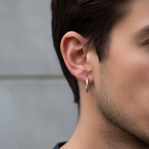 May include: A close-up of a silver hoop earring. The earring is circular and hangs from the ear. The background is a blurred gray. The earring is a simple, minimalist design.