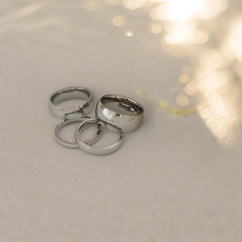 A group of silver wedding rings is arranged on a light-colored surface, with a blurred background suggesting a natural setting.