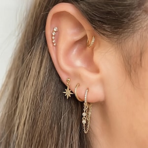 May include: Close-up of an ear adorned with multiple gold earrings. A helix piercing features a series of small, sparkling stones. Other earrings include a starburst stud, a hoop, and a chain drop earring, all in gold.
