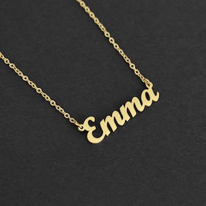 May include: A gold-colored necklace with the name "Emma" in a cursive font. The chain is delicate, and the pendant is centered. The necklace is displayed against a dark background, highlighting the gold color.