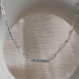 May include: A silver-toned paperclip chain necklace featuring three rectangular metal bars. One bar is engraved with the name Megan, another with the letters AOE and a heart symbol, and the bottom bar displays the date 06.30.2022 in black text.