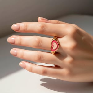 May include: A gold-toned ring featuring a heart-shaped design with a vibrant pink enamel border surrounding a light pink center. The ring has a smooth, polished finish and a tapered band that sits comfortably on the finger, showcasing a clean aesthetic.