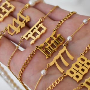 A hand holding multiple gold necklaces with various pendants, including numbers and pearls.