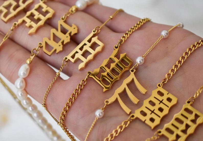 A hand holding multiple gold necklaces with various pendants, including numbers and pearls.