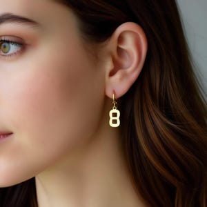 May include: A close-up of a gold-colored earring shaped like the number eight. The earring is hanging from a small hoop and is worn on a person's ear. The background is blurred, focusing on the jewelry.