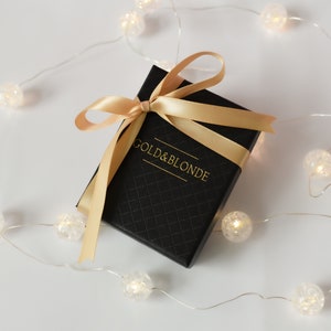 May include: A black gift box with a gold ribbon and the text "GOLD & BLONDE" on the top. The box is surrounded by a string of white lights.